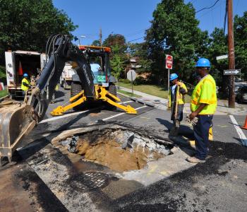 Two workers making a water main break repair with a excavator nearby